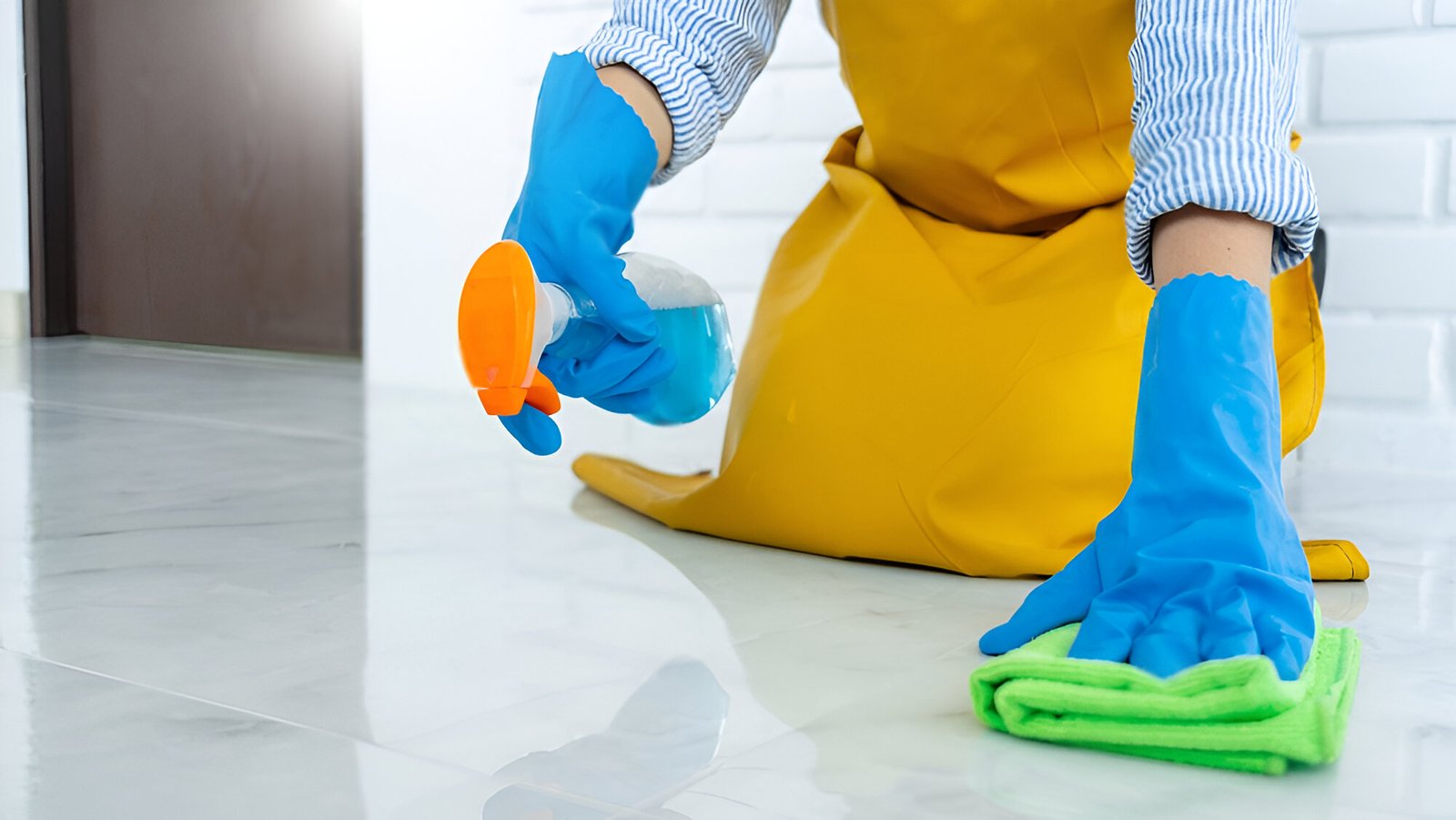 Person wearing blue gloves and yellow apron cleaning a tiled floor with a green microfiber cloth and blue disinfectant spray bottle.